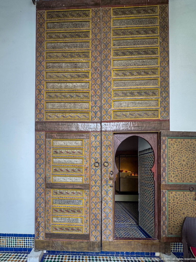 Decorated wooden wall and doorway with geometric painted panels, small arched opening, patterned floor, and blue tile border.