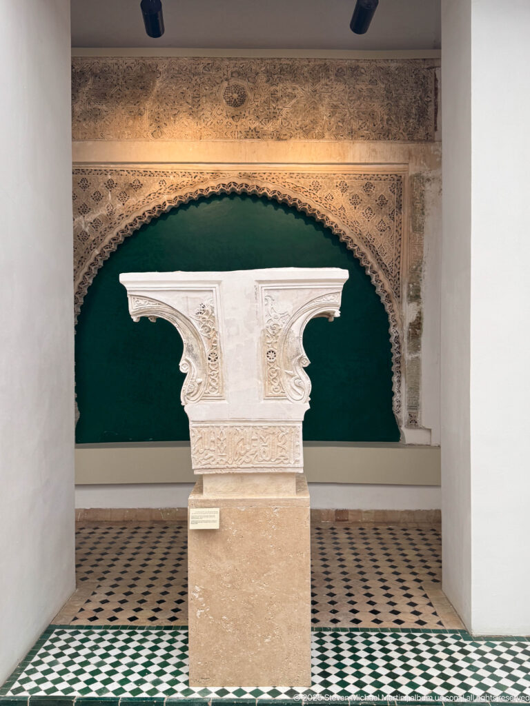 Pale carved capital-like architectural fragment on a pedestal before a broad scalloped arch with relief ornament.