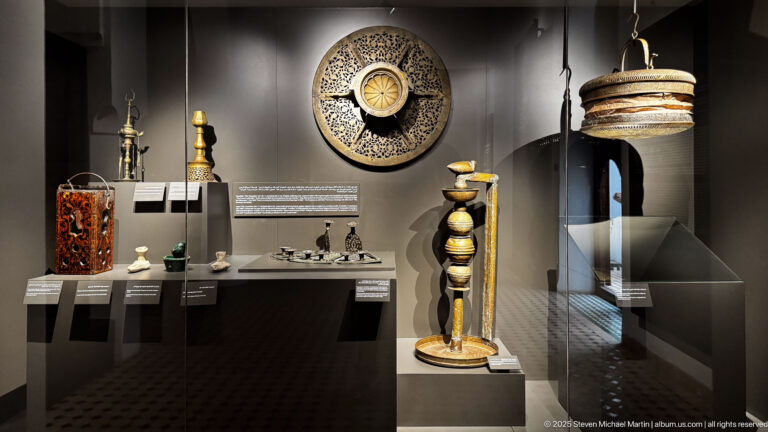 Dark museum display with candlestick forms, pierced metal objects, a circular disk, tall stand, and suspended tiered lamp.