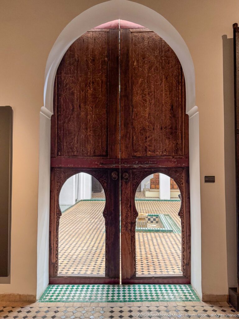 Monumental reddish carved wooden doors with two symmetrical lower arches opening onto a brightly tiled gallery floor.