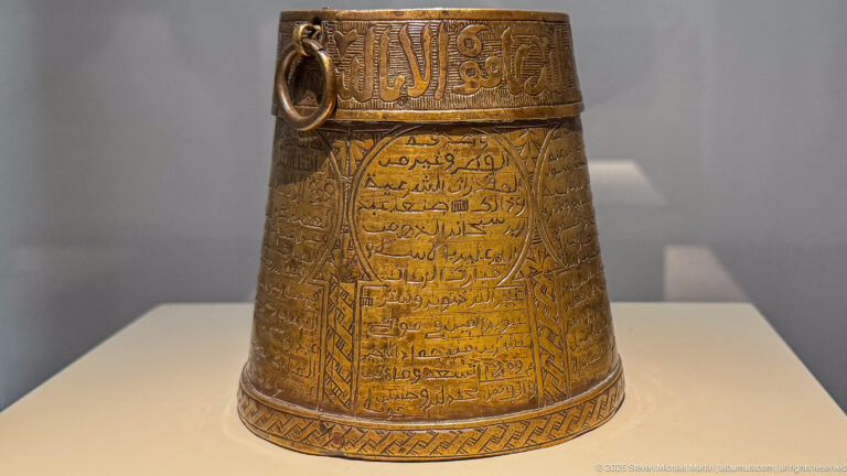 Tapered golden-brown metal vessel with Arabic inscription panels, geometric borders, and ring handles near the rim.