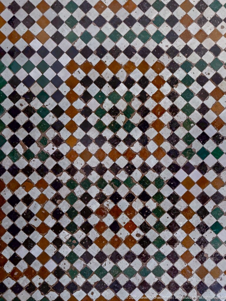 Close-up of worn multicolored diamond-pattern floor tiles in green, ochre, white, and dark tones.