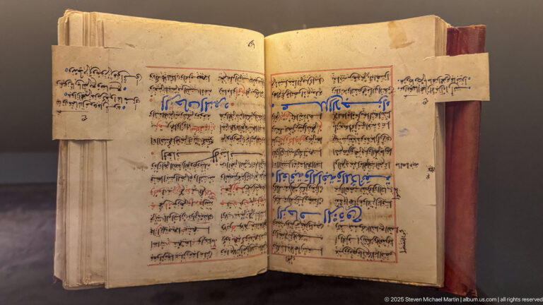 Open Arabic manuscript filled with dark script, red and blue notation, and narrow paper extensions projecting from the page margins.