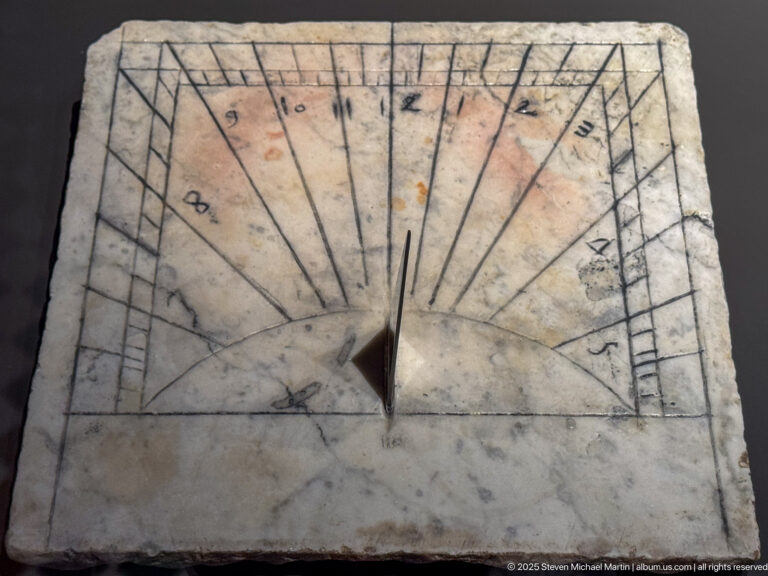 Square pale stone sundial with radiating incised hour lines, small markings, and a triangular metal gnomon at center.