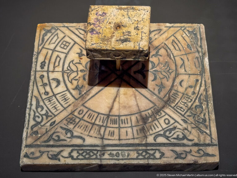 Small square sundial-like instrument with incised radial scale, numerals, decorative motifs, and a raised rectangular block at center.
