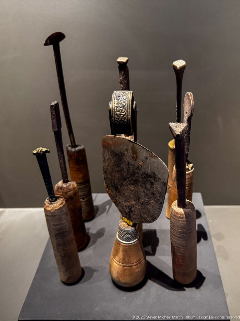Small museum display of cylindrical inkwells or pen holders, reed-like writing implements, and a dark central metal object.