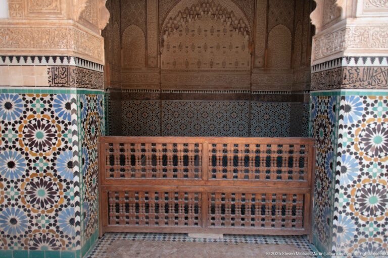 Wooden lattice barrier before a pointed decorated recess, flanked by colorful zellij and carved stucco walls.
