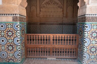Wooden lattice barrier before a pointed decorated recess, flanked by colorful zellij and carved stucco walls.