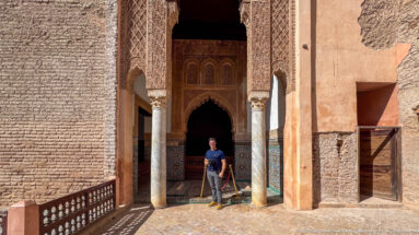 Visitor standing between columns of a carved and tiled portal leading into a shadowed Saadian tomb chamber.