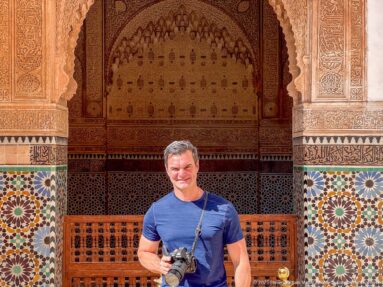 Visitor holding a camera before carved stucco, colorful zellij, and a wooden lattice screen inside the Saadian Tombs.