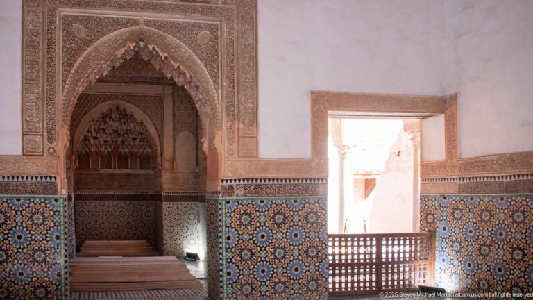 Pointed carved niche and colorful zellij beside a bright courtyard opening with wooden lattice railings.