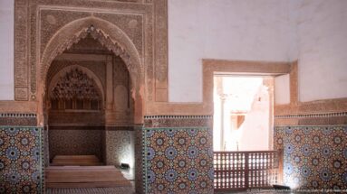 Pointed carved niche and colorful zellij beside a bright courtyard opening with wooden lattice railings.