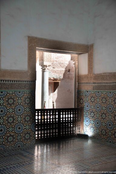 Geometric zellij wall and patterned floor around a bright rectangular opening with a dark wooden lattice railing.