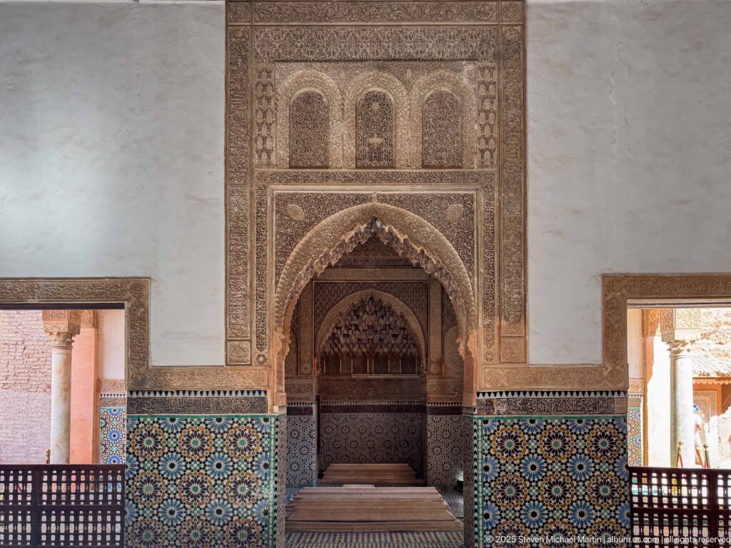 Symmetrical chamber wall with central carved pointed recess, colorful zellij, side openings, and wooden railings.