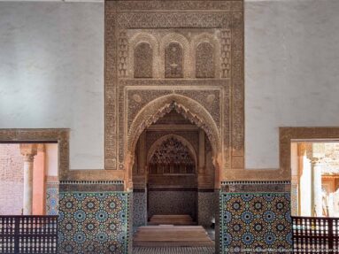 Symmetrical chamber wall with central carved pointed recess, colorful zellij, side openings, and wooden railings.