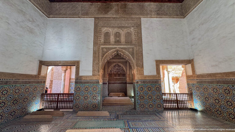 Wide symmetrical room with central carved recess, zellij-covered lower walls, patterned floor, and bright side openings.