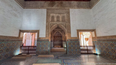 Wide symmetrical room with central carved recess, zellij-covered lower walls, patterned floor, and bright side openings.