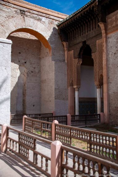 Shaded carved portico with slender columns and timber eave behind wooden courtyard balustrades and weathered walls.