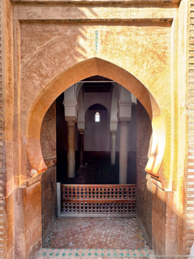 Large sunlit horseshoe arch opening into a dim chamber with pale columns, smaller arches, and a wooden lattice barrier.