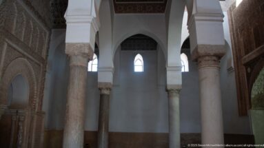 Marble columns and white arches rise between carved stucco walls inside an ornate funerary chamber.