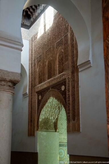 Carved stucco arch with dense geometric ornament opens toward a bright muqarnas-decorated space.