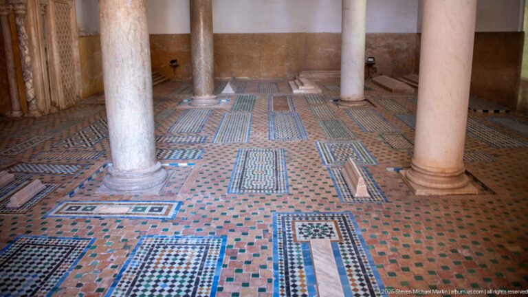 Geometric zellij paving surrounds several rectangular grave markers between marble columns.