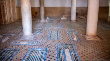 Geometric zellij paving surrounds several rectangular grave markers between marble columns.
