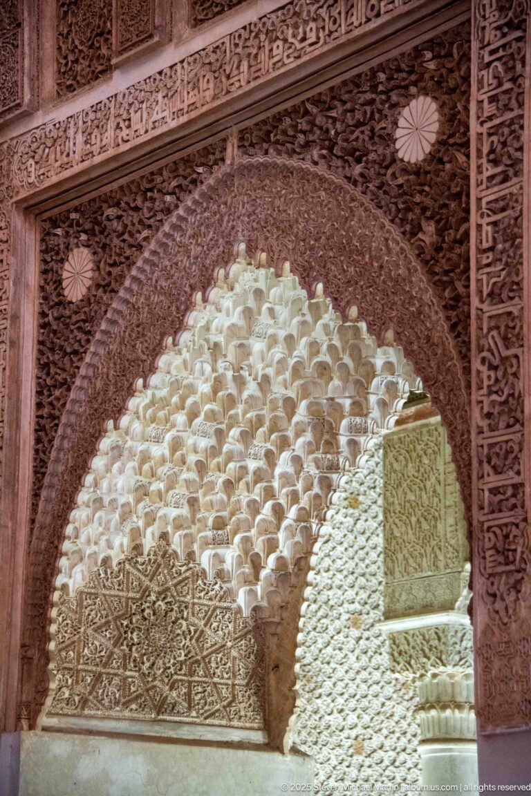 Close view of pale muqarnas cells framed by a dark, densely carved stucco arch.
