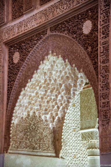 Close view of pale muqarnas cells framed by a dark, densely carved stucco arch.