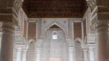 Frontal interior view of marble columns, carved stucco arches, blue-green zellij, and dark wooden ceiling.