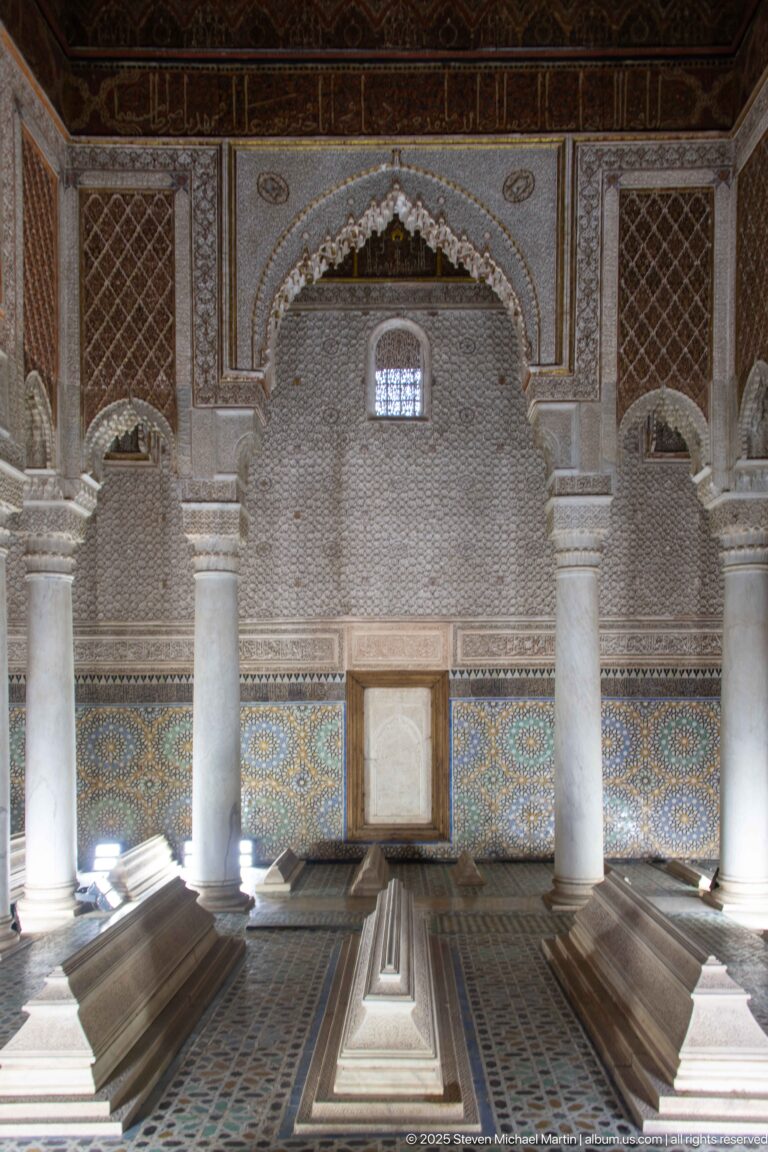 Raised tombs align toward a carved arched wall with zellij dado and a small high window.
