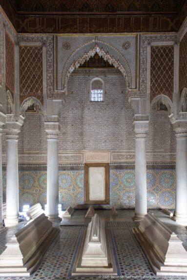 Raised tombs align toward a carved arched wall with zellij dado and a small high window.