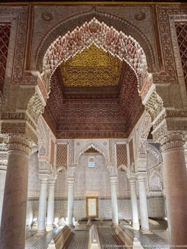Upward view through a richly carved arch toward a dark wooden ceiling and marble columns.