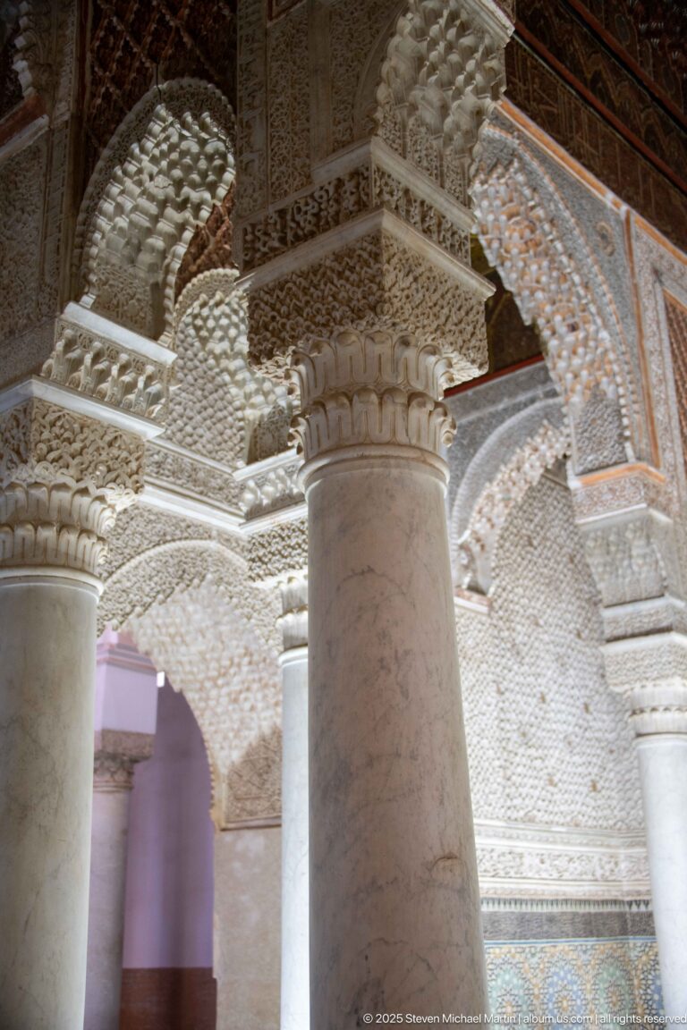 Close oblique view of a marble column meeting a deeply carved stucco capital and arches.