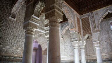 Oblique view of marble columns, carved stucco arches, zellij wall band, and dark ceiling.