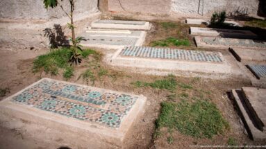Foreground grave with green rosette-patterned mosaic beside other tiled rectangular graves on grass and bare earth.
