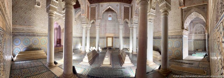 Long view through a columned tomb chamber with low graves, carved arches, tiled walls, and patterned floor.