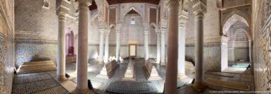 Long view through a columned tomb chamber with low graves, carved arches, tiled walls, and patterned floor.