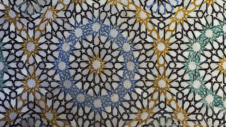 Close-up of polychrome zellij mosaic with interlaced black lines, starbursts, and blue, green, ochre, and white pieces.