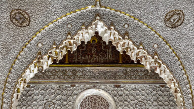 Close-up of a muqarnas-edged arch, carved stucco, and dark Arabic inscription panel above the opening.