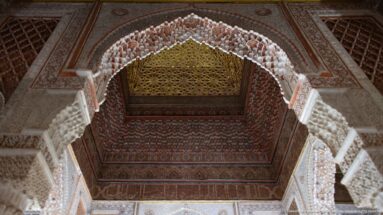 Upward view through carved white stucco arch toward a painted geometric wooden ceiling panel.