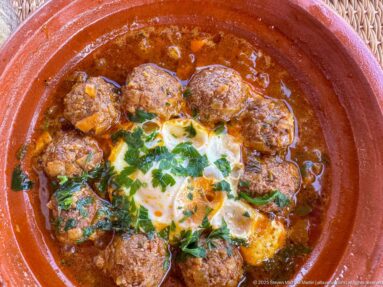 Earthenware dish of meatballs in red sauce topped with egg and chopped green herbs.