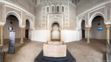 Ornate chamber with carved stucco walls, horseshoe arches, and a rectangular stone feature centered in the foreground.