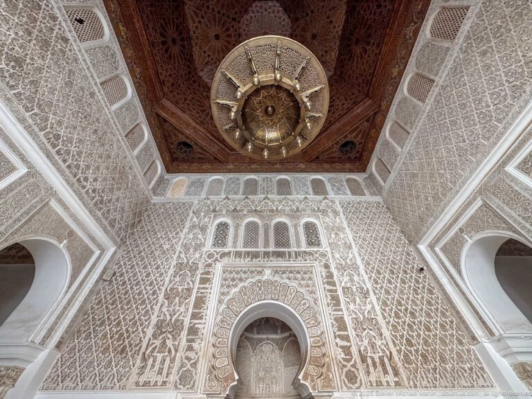 Large hanging metal lamp beneath decorated wooden ceiling and carved stucco walls with small arches.