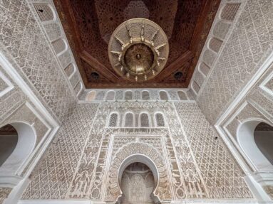 Large hanging metal lamp beneath decorated wooden ceiling and carved stucco walls with small arches.