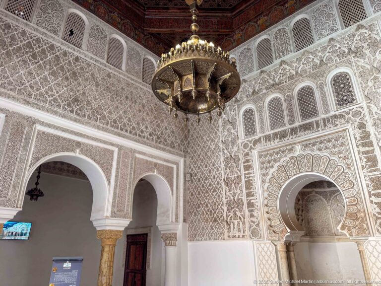 Corner of ornate chamber with hanging metal lamp, horseshoe arches, carved stucco, niche, and wooden door.