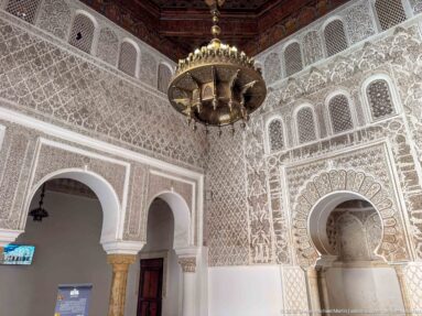 Corner of ornate chamber with hanging metal lamp, horseshoe arches, carved stucco, niche, and wooden door.