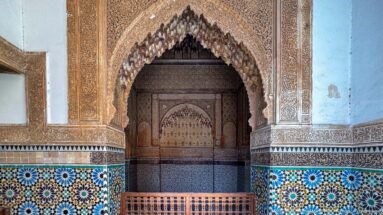 Pointed carved-stucco recess above colorful star-patterned zellij and a low wooden screen in a Saadian tomb chamber.