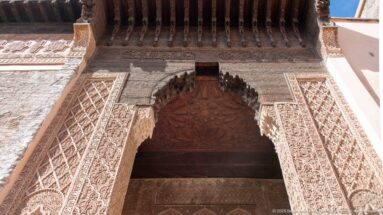 Upward view of an ornate carved-stucco portal and scalloped arch beneath a dark projecting wooden eave.