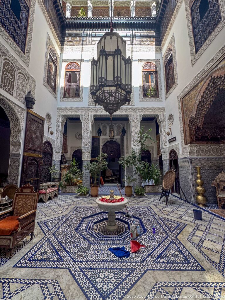 Ornate riad courtyard with central fountain, blue-and-white tile, carved plaster, seating, plants, and a large hanging lantern.